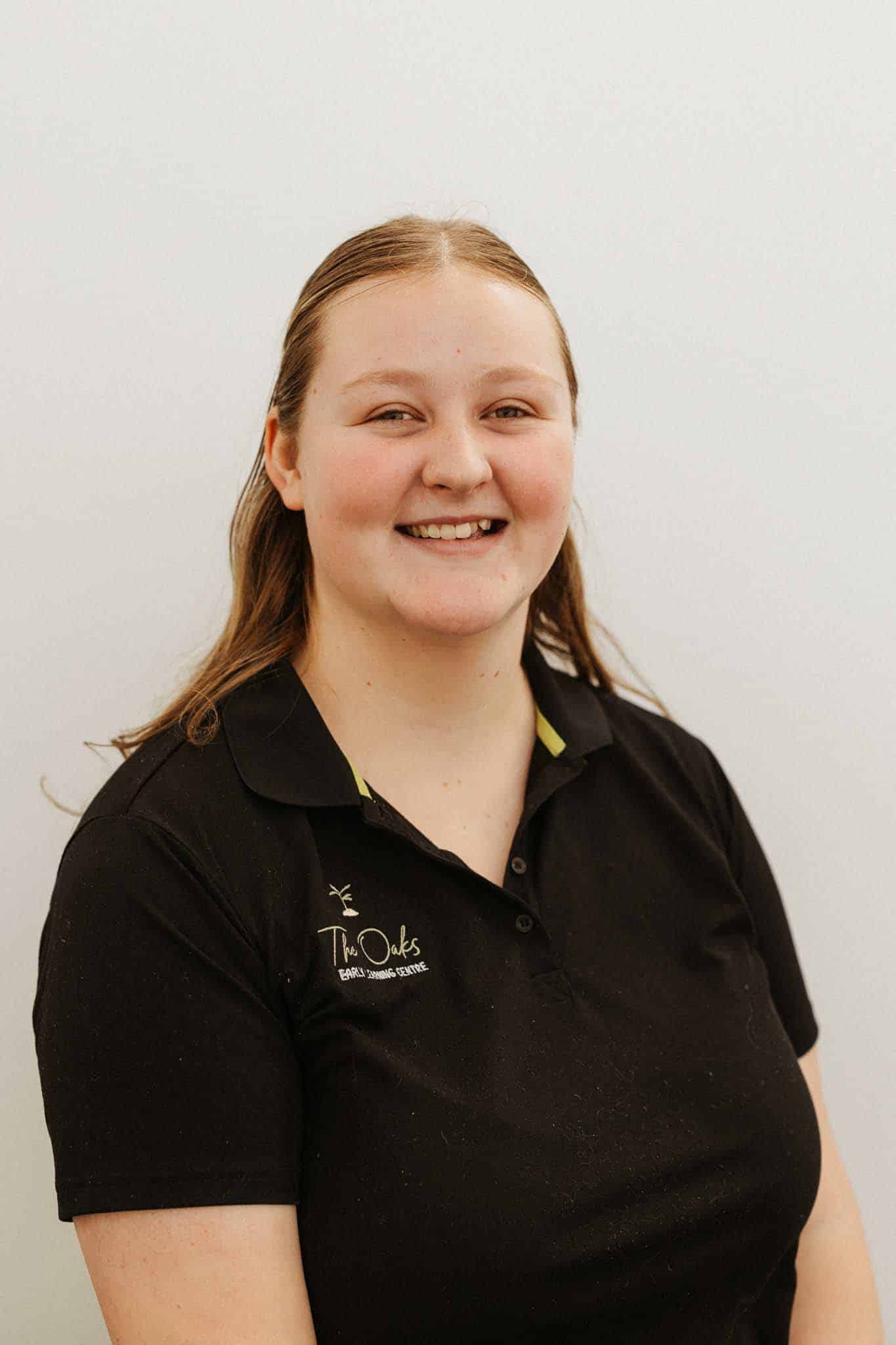 A young woman with long brown hair wearing a black polo shirt with "The Oaks" logo, smiling against a plain light background.