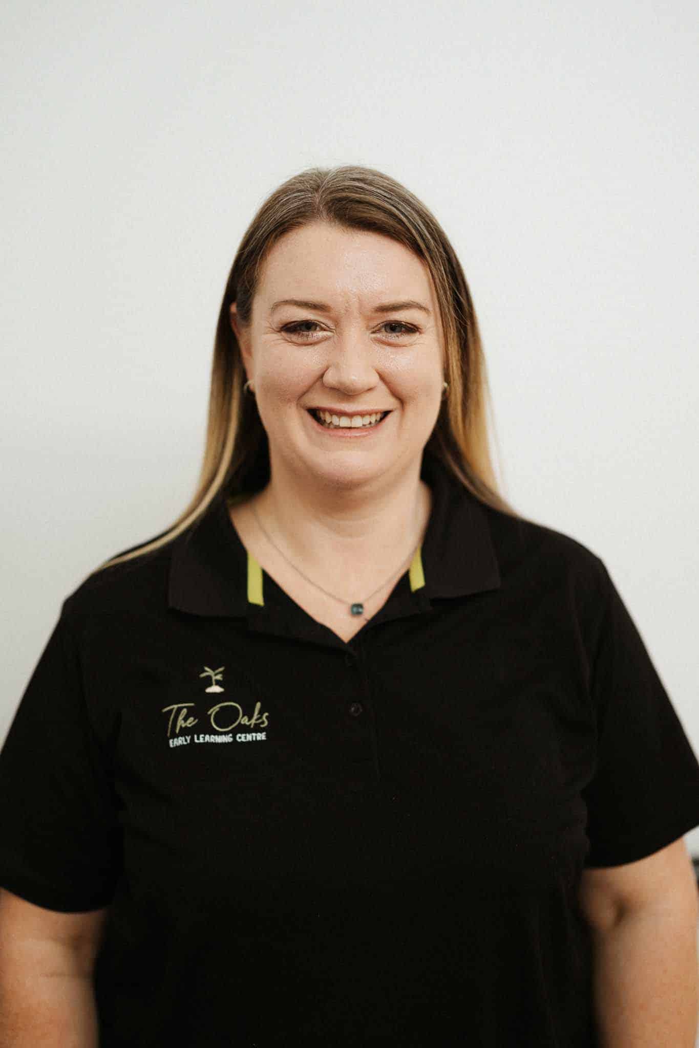 A woman with straight, light brown hair smiles at the camera while wearing a black polo shirt with "The Oaks Early Learning Centre" embroidered on it. Plain light background.