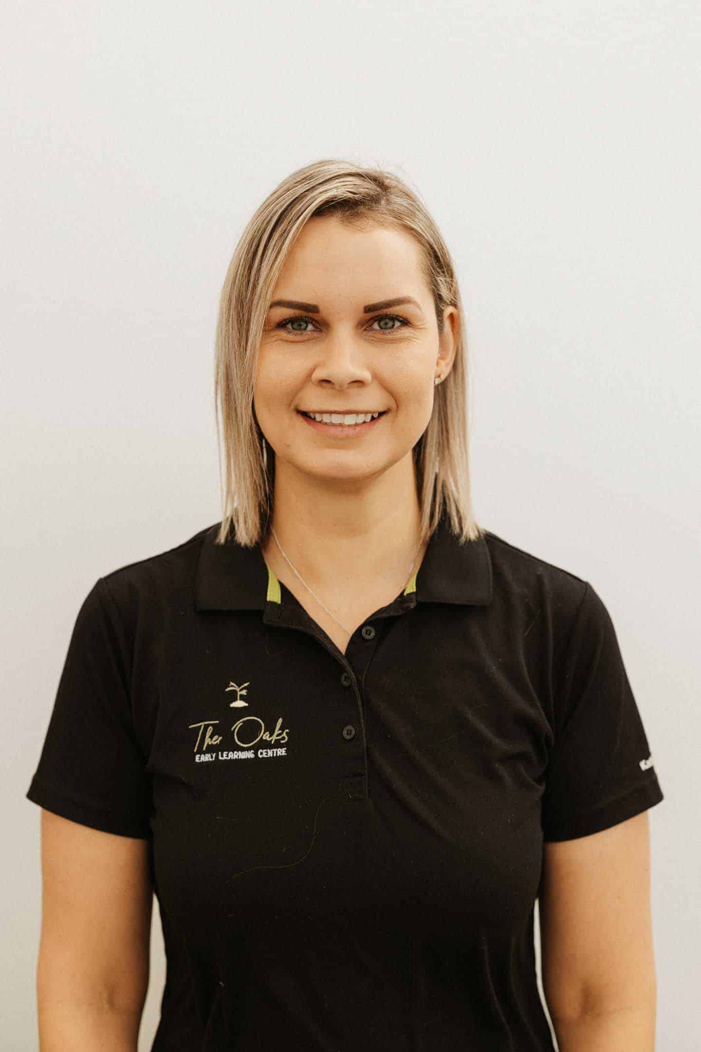 A woman with straight, shoulder-length blonde hair is wearing a black polo shirt with "The Oaks Early Learning Centre" logo, standing against a plain light background.