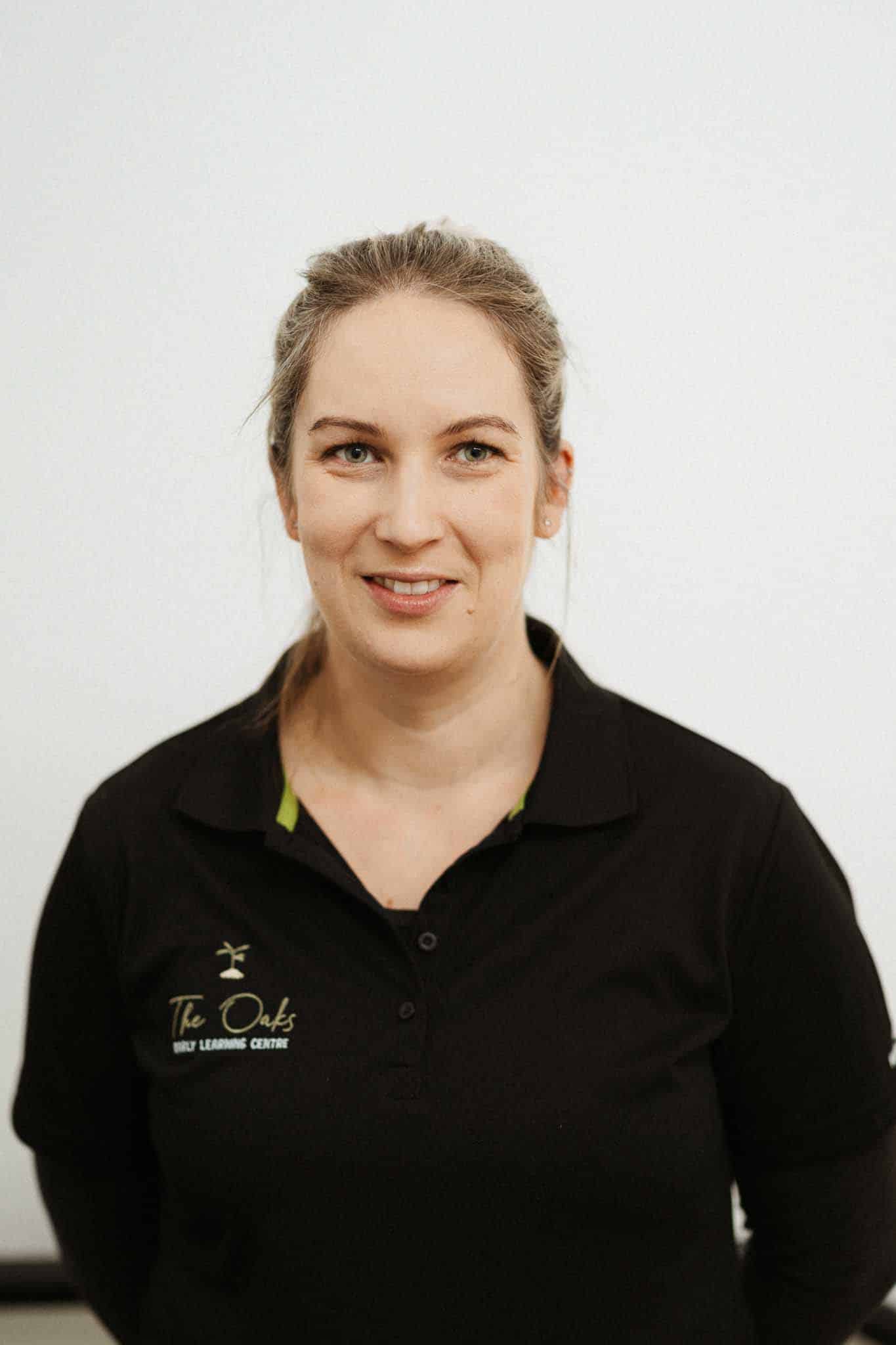 A woman with light hair tied back, wearing a black collared shirt with "The Oaks Early Learning Center" logo, stands in front of a plain white background.