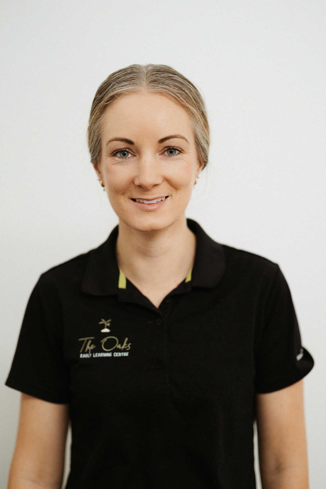 A woman with light hair, wearing a black collared shirt with "The Oaks Early Learning Centre" logo, stands against a plain white background.