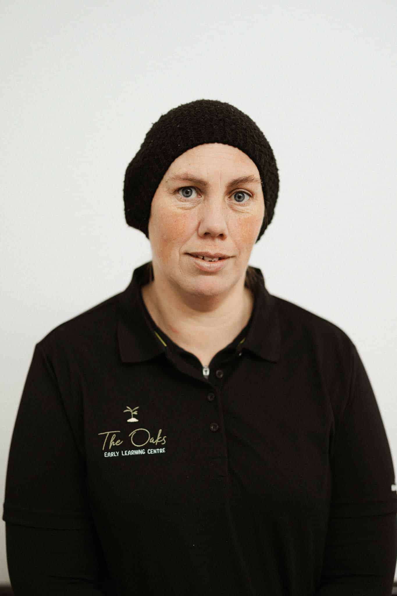 A woman wearing a black head covering and a black "The Oaks Early Learning Centre" polo shirt stands against a plain white background.