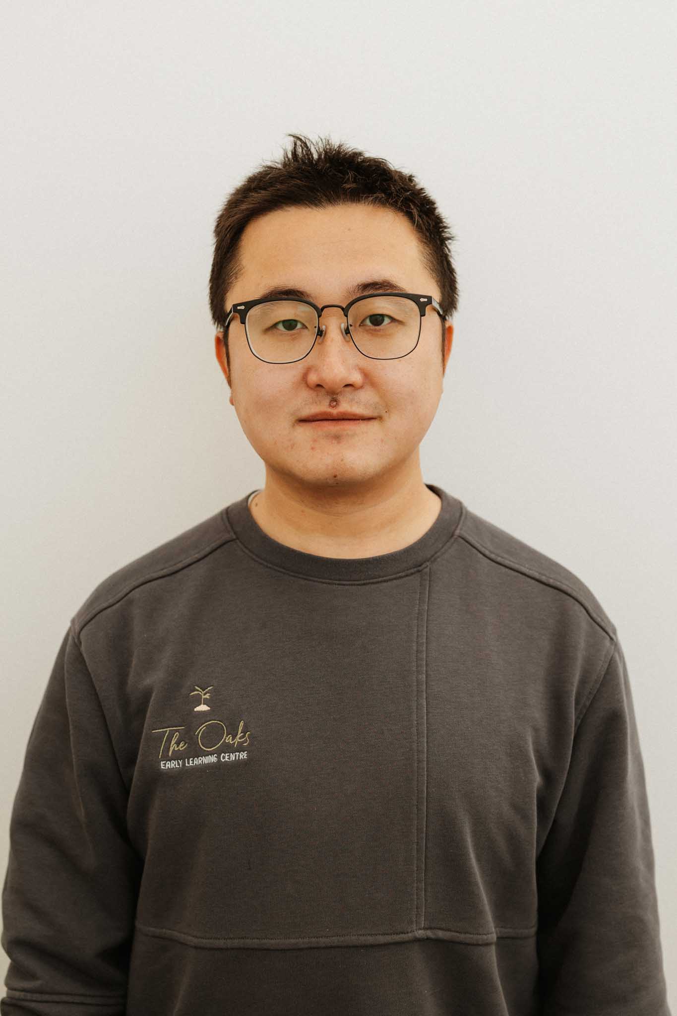 A person with short dark hair and glasses wearing a grey sweatshirt that reads "The Oaks Early Learning Centre" stands against a plain light background.
