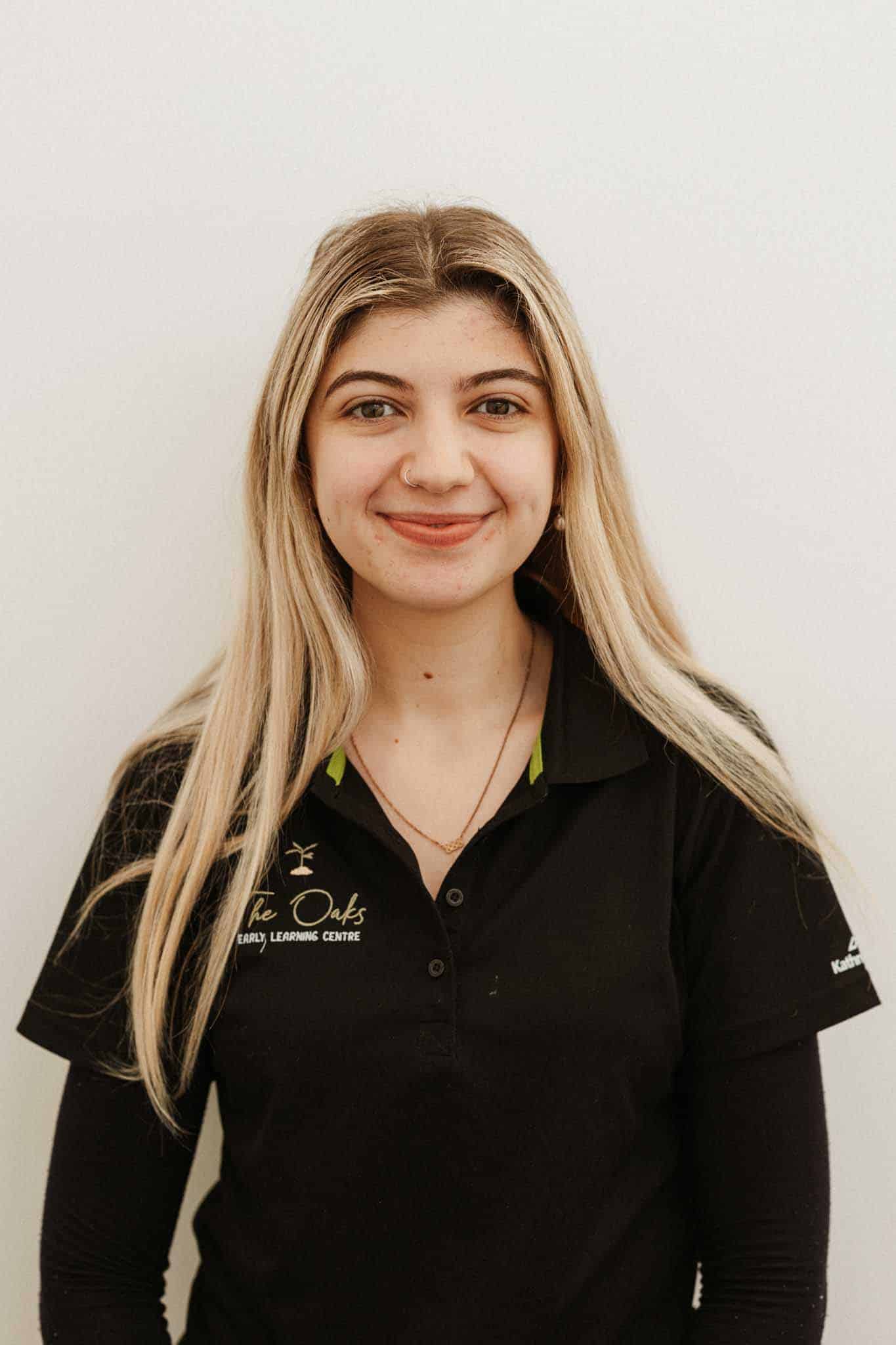 A young woman with long blonde hair stands against a plain background, wearing a black polo shirt with a logo that reads "The Oaks Early Learning Centre.