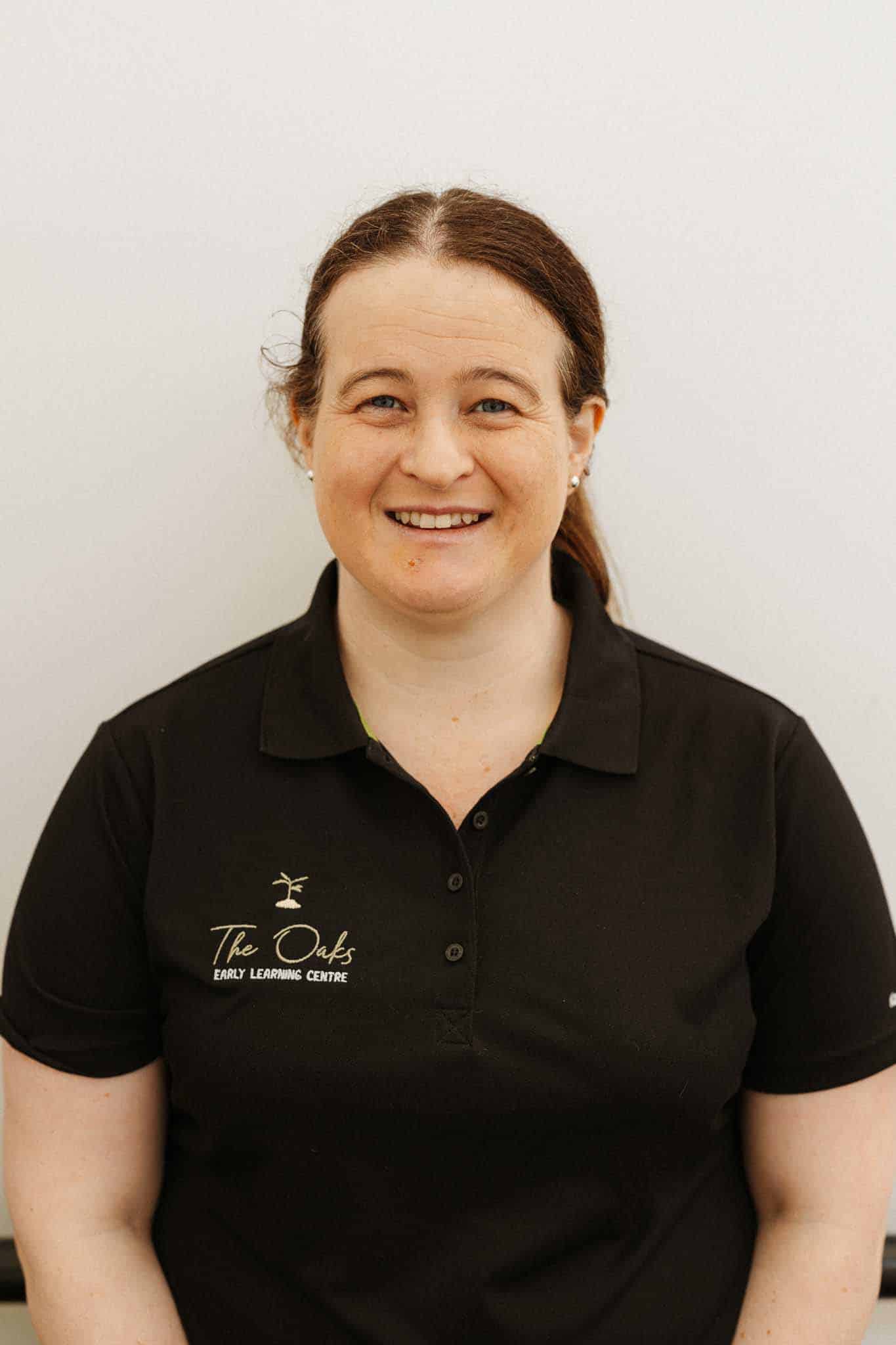 A woman with long brown hair, tied back, wearing a black polo shirt with "The Oaks Early Learning Centre" logo, stands against a plain light-colored background, smiling at the camera.