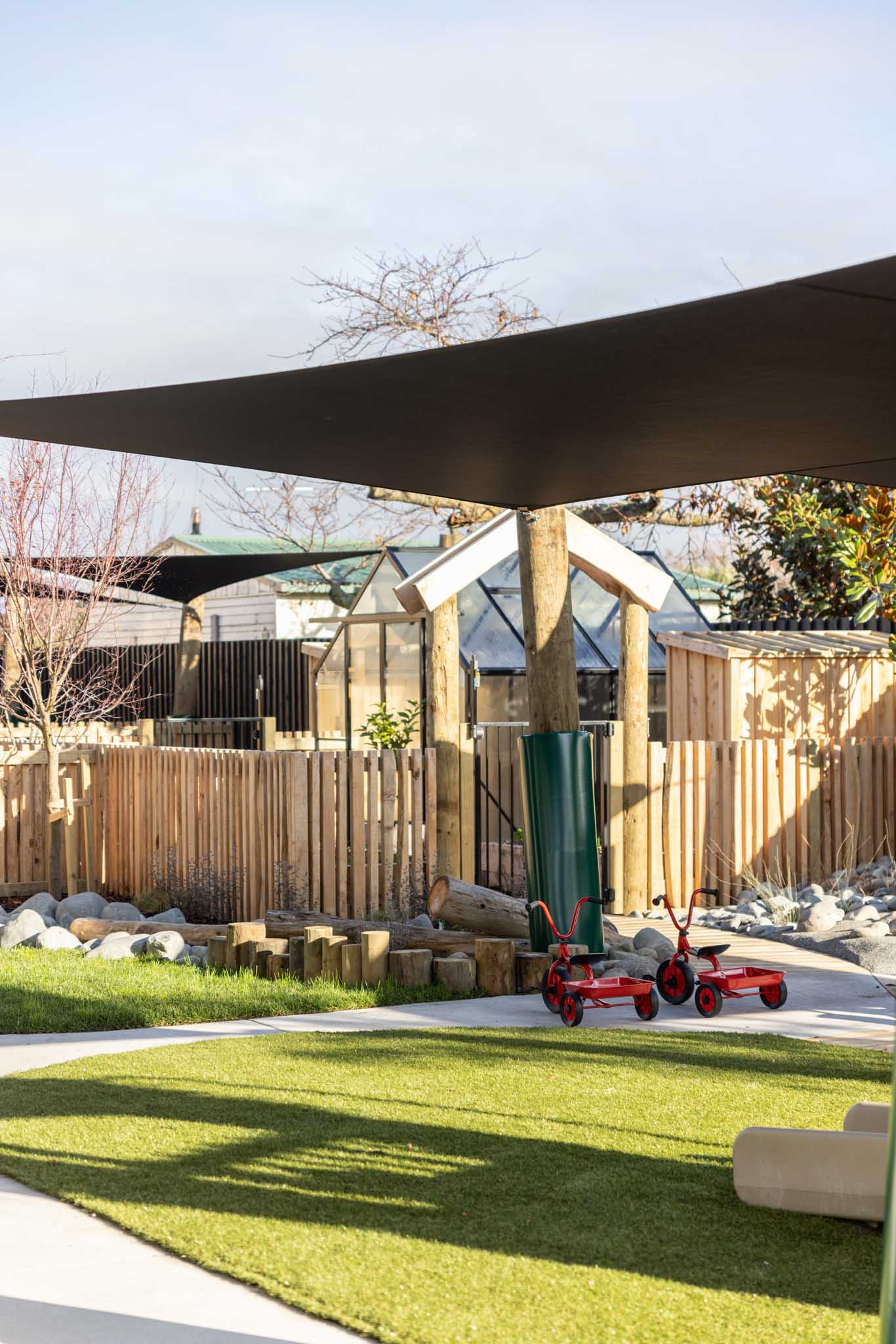 Outdoor playground area with artificial grass, wooden fence, small greenhouse, and two red tricycles under shade sails on a sunny day.