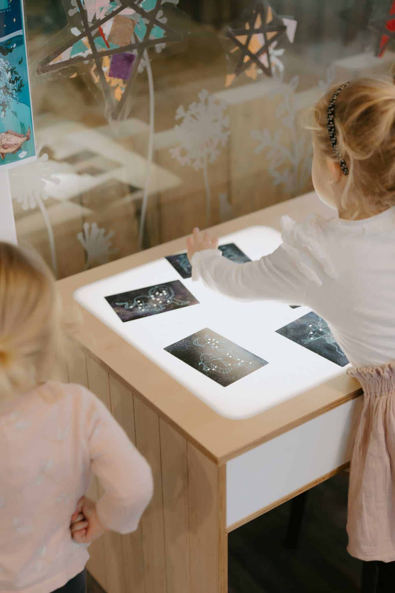 Two young children interact with constellation cards on a light table in a classroom or learning environment.