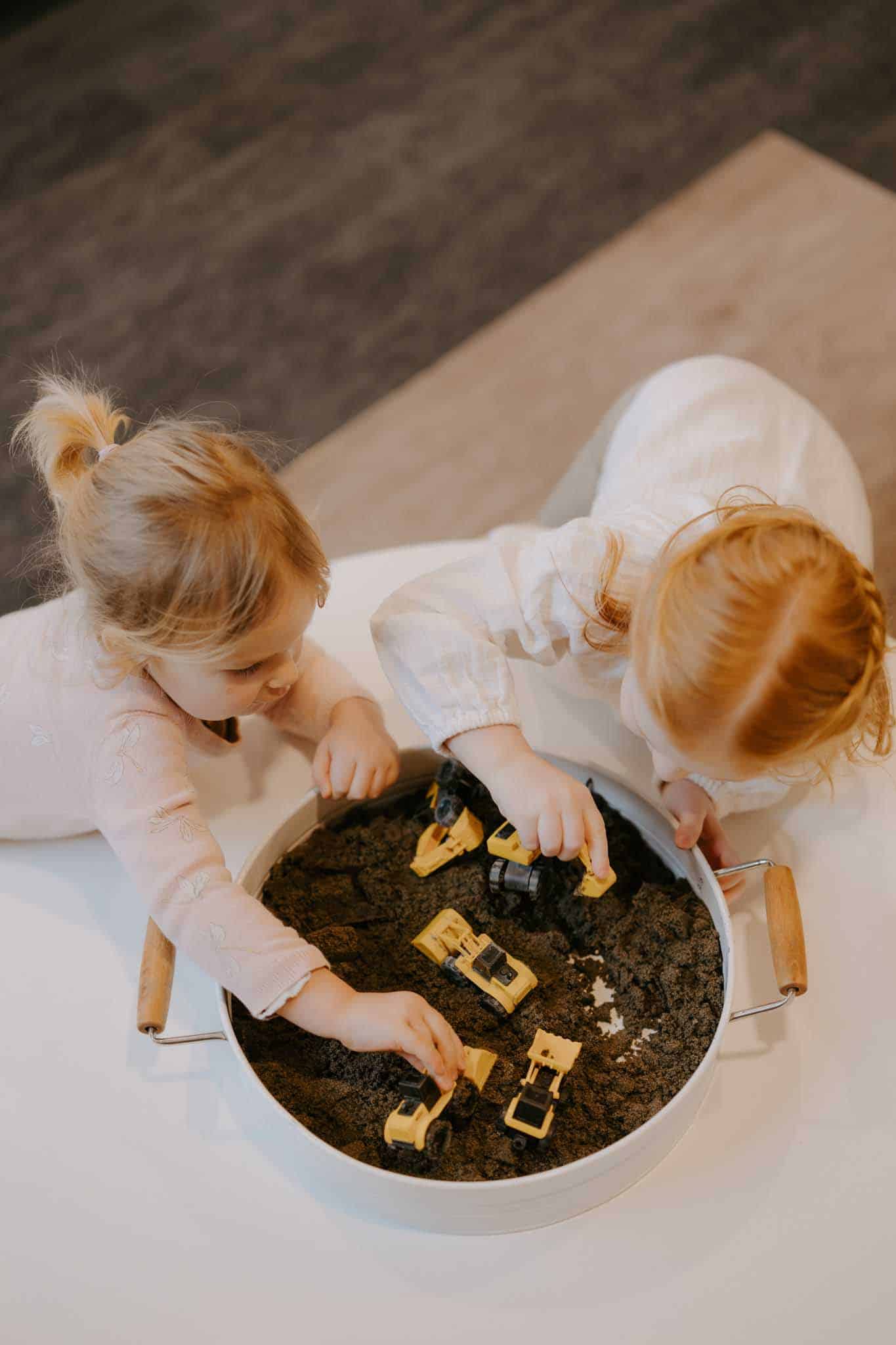Two young children are playing with small yellow construction vehicles in a round container filled with dirt.