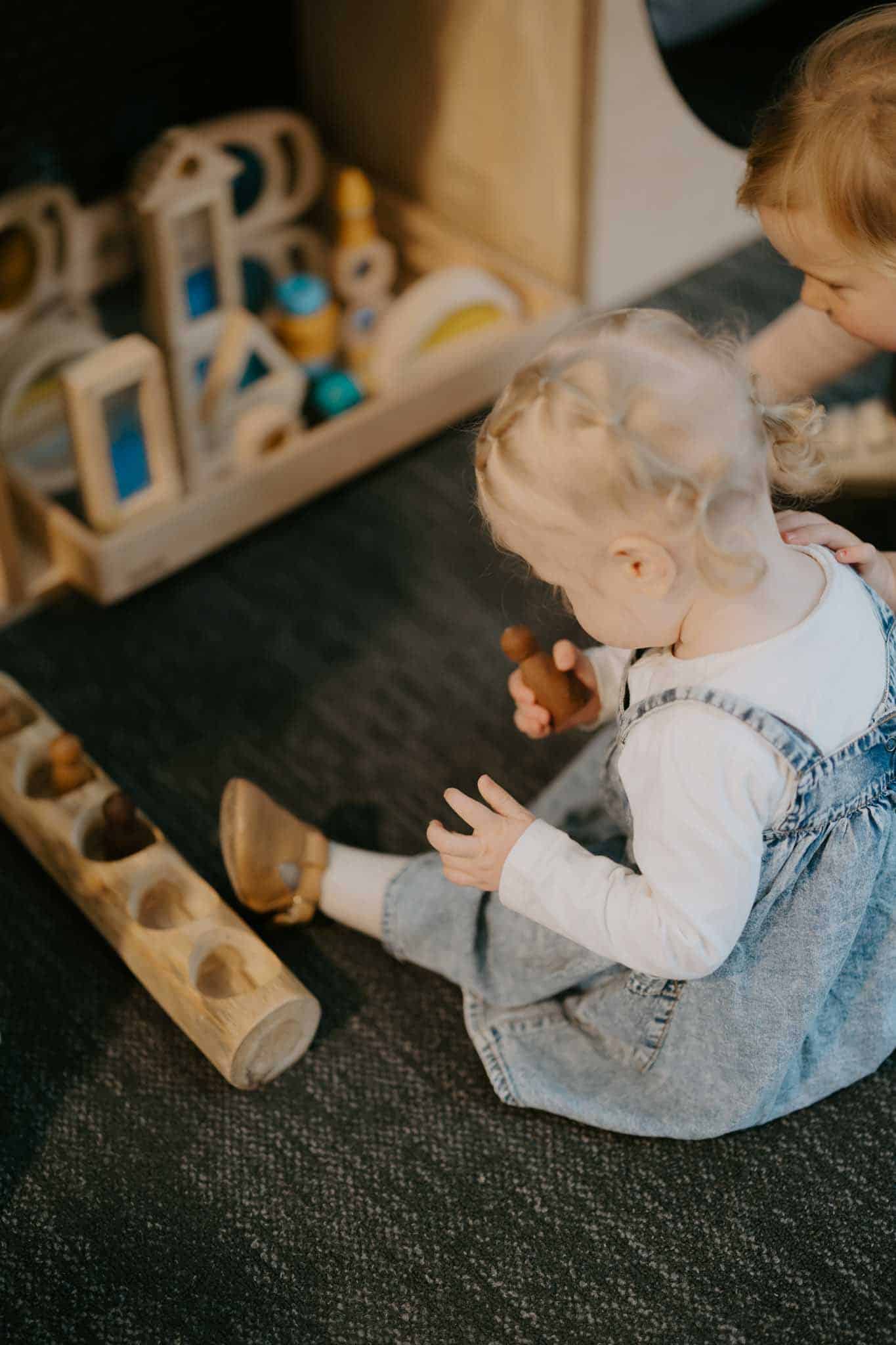 Two young children sit on the floor playing with wooden toys and building blocks.