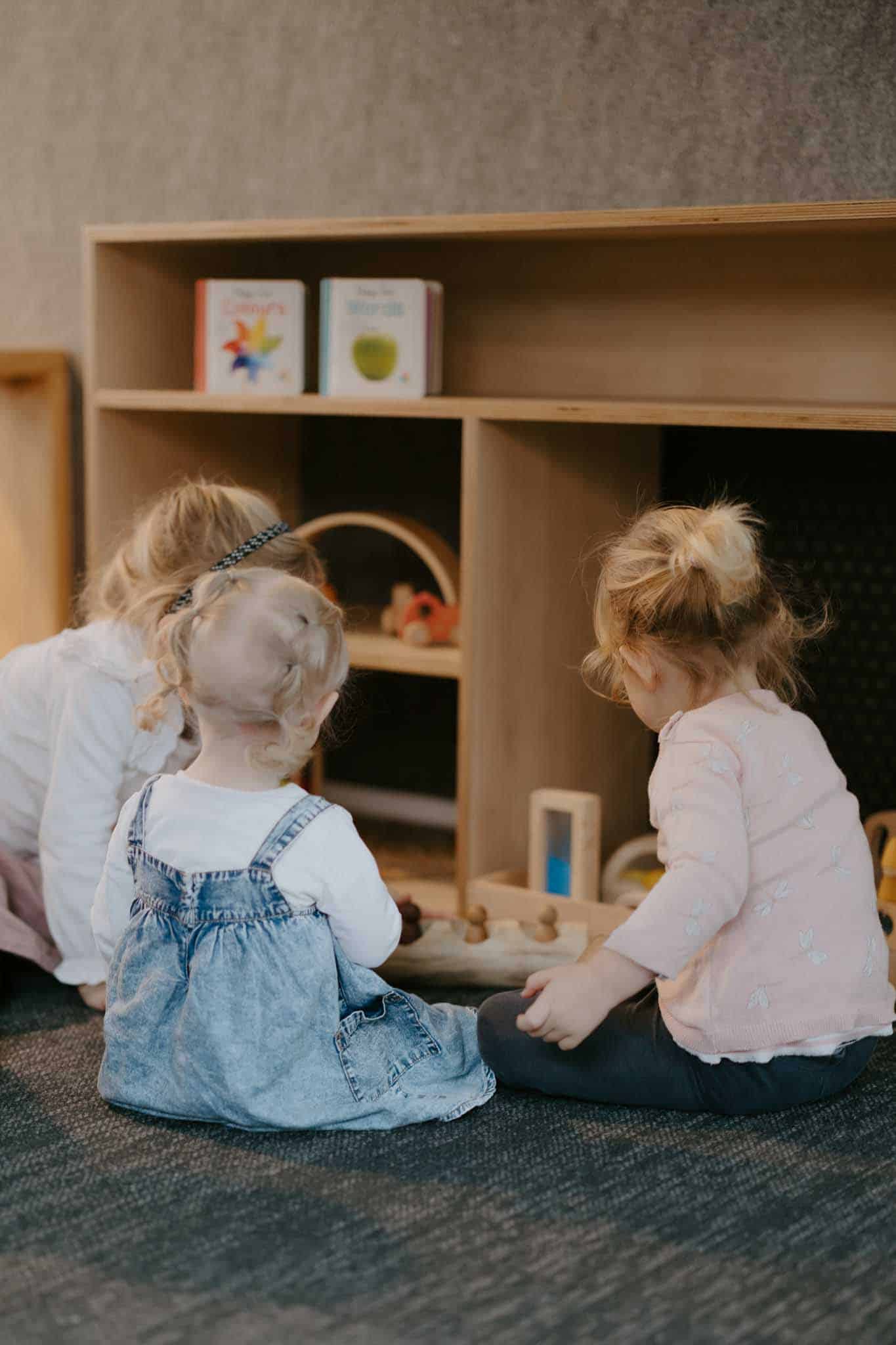 Three young children sit on the floor playing with wooden toys in front of a low bookshelf with books displayed on top.