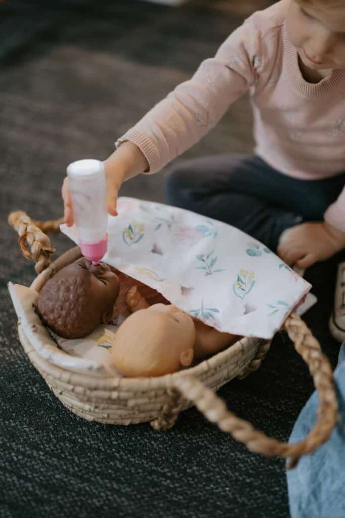 A child holds a toy bottle to the mouth of a baby doll lying in a woven basket beside another doll, partially covered by a floral blanket.