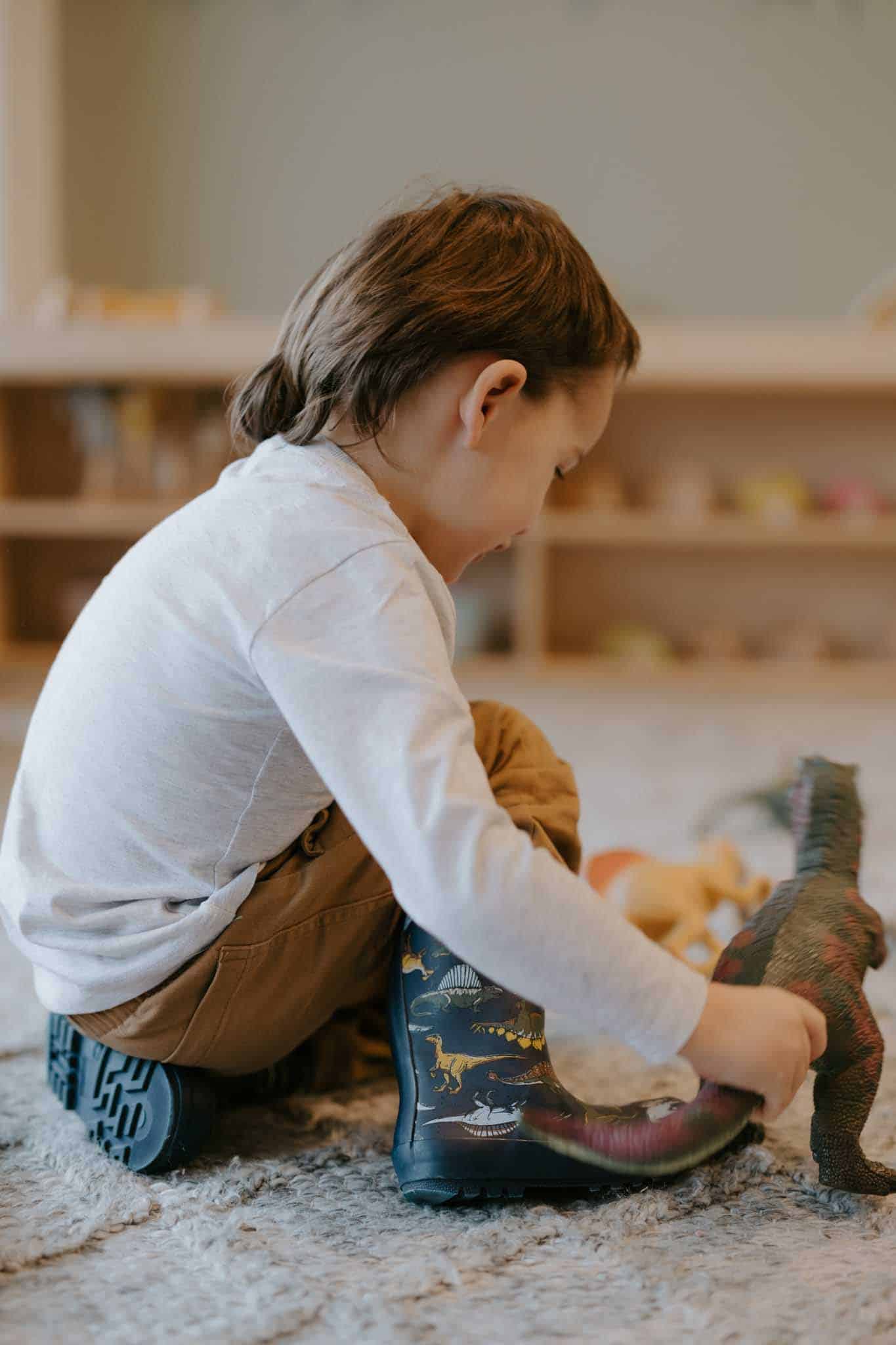 A child with brown hair sits on the floor, playing with dinosaur toys. The child is wearing a light gray shirt, brown pants, and blue rain boots with dinosaur designs.