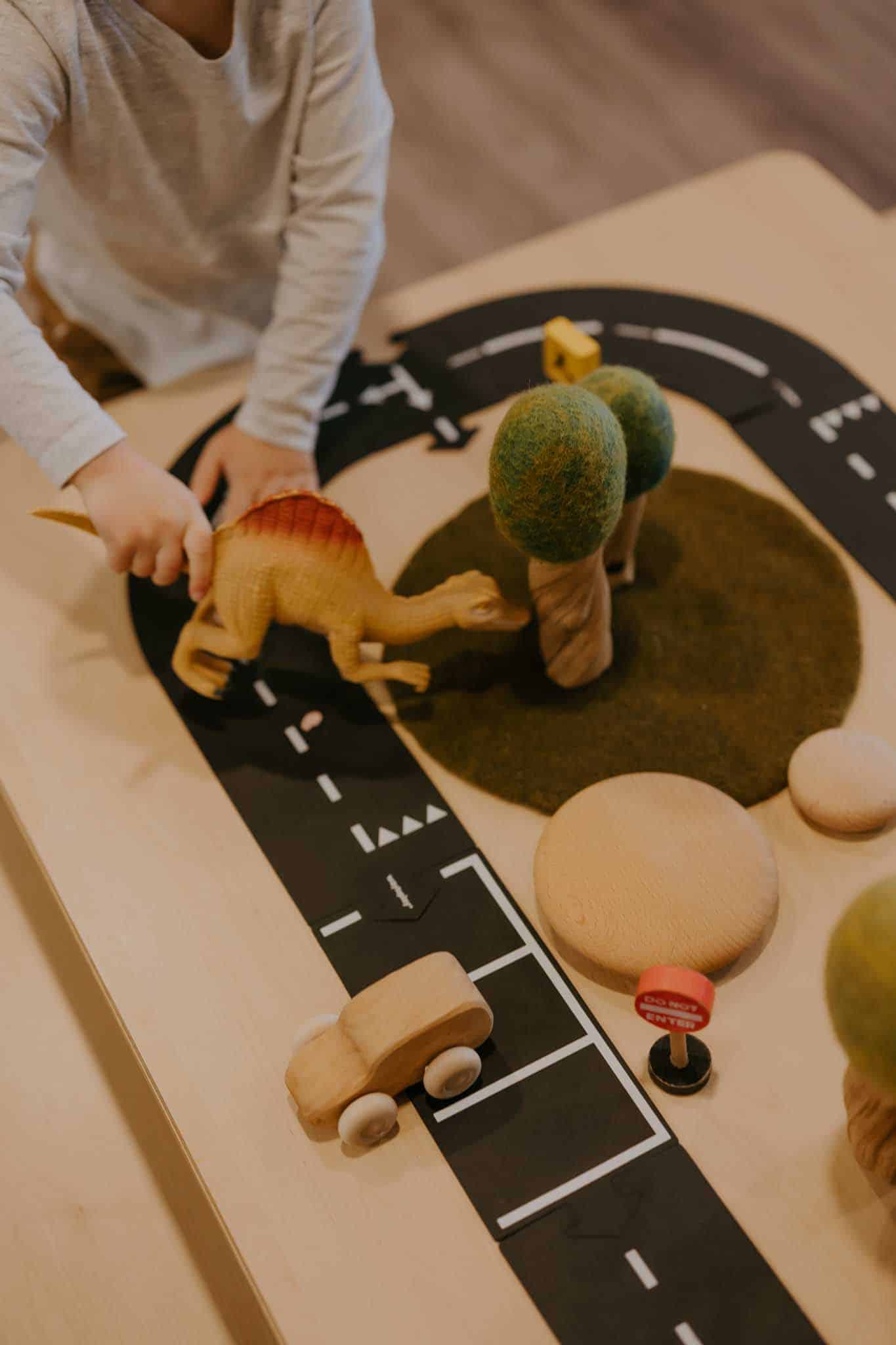 A child plays with a toy dinosaur on a tabletop road scene with wooden trees, a wooden car, and various small objects.