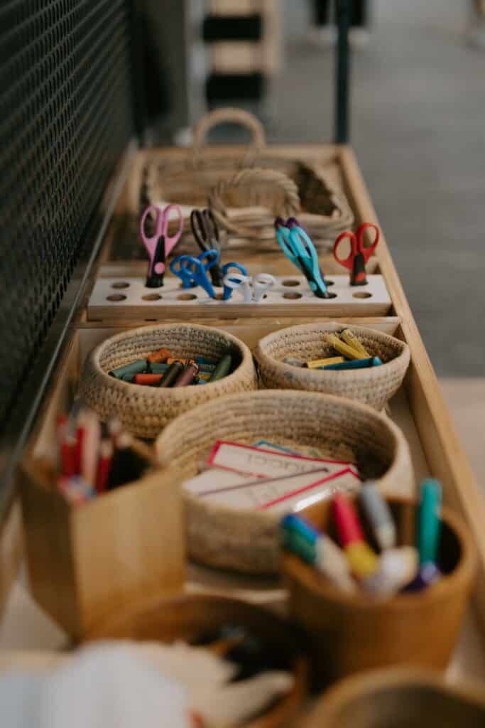 Assorted baskets and containers holding scissors, colored pencils, markers, and other art supplies are arranged on a wooden table.
