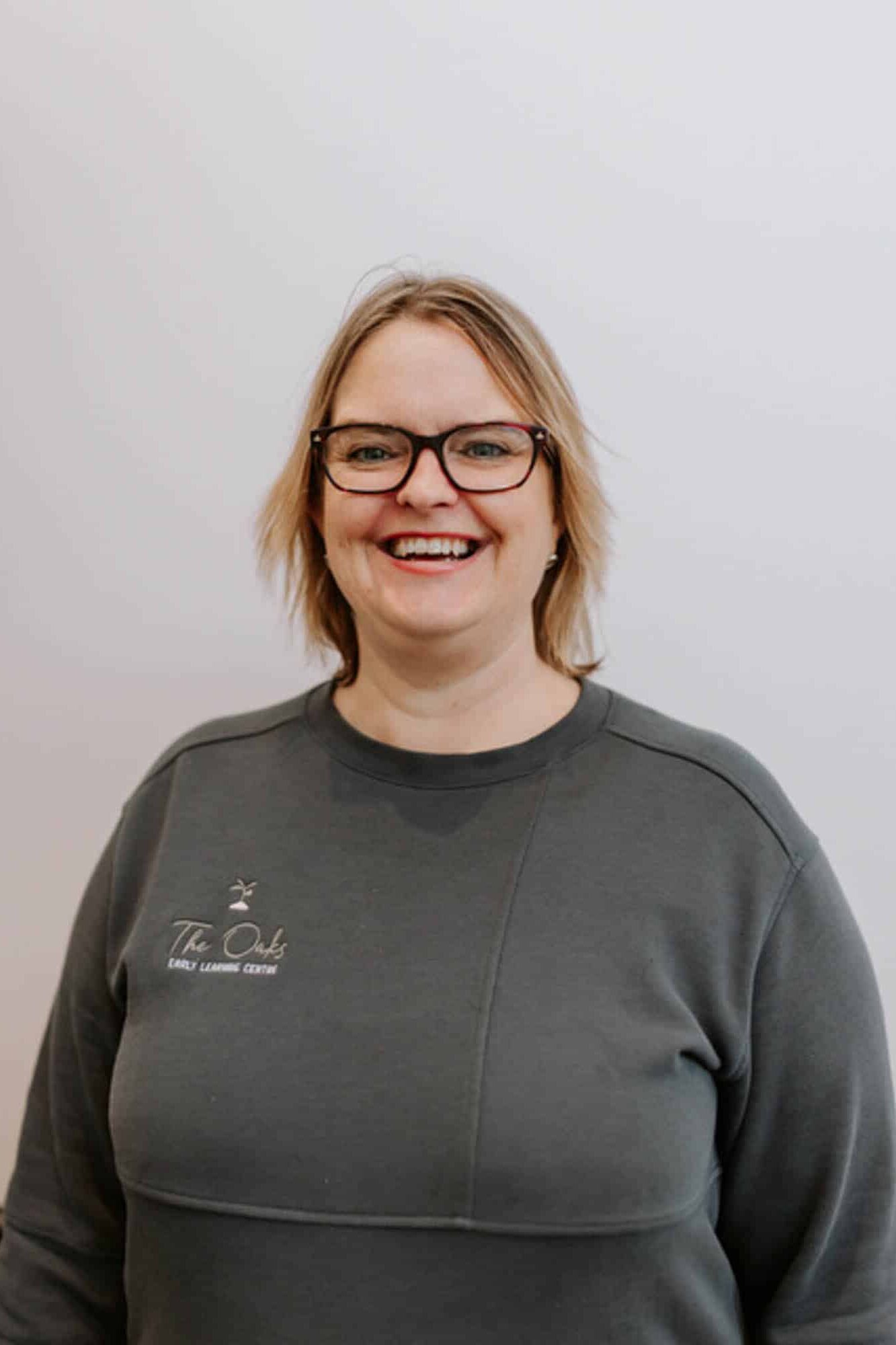 A woman with glasses, shoulder-length blonde hair, and a grey sweatshirt stands in front of a plain light background, smiling at the camera.