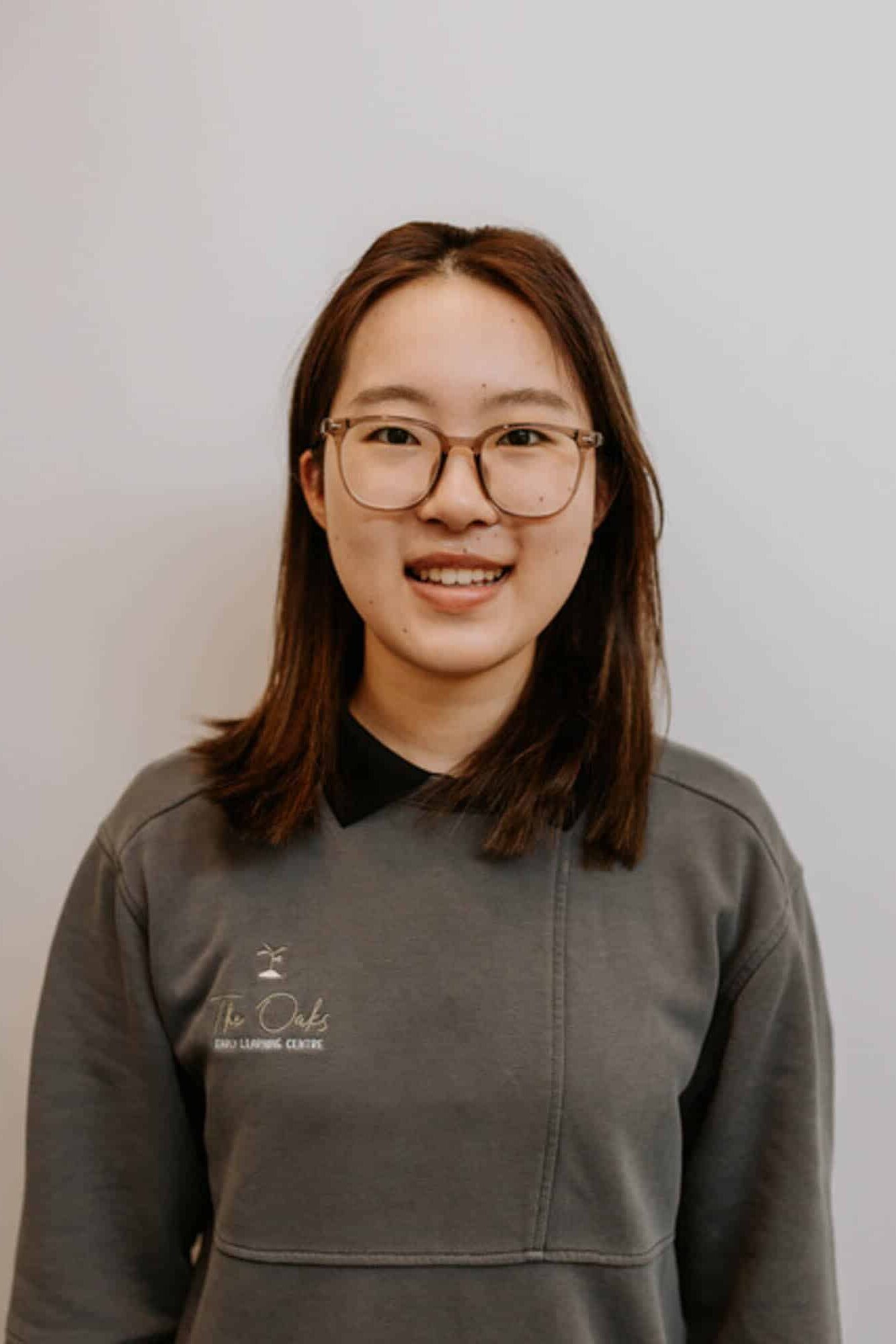 A woman with straight brown hair and glasses is standing against a plain white background, wearing a grey sweatshirt with a small logo on the left chest.