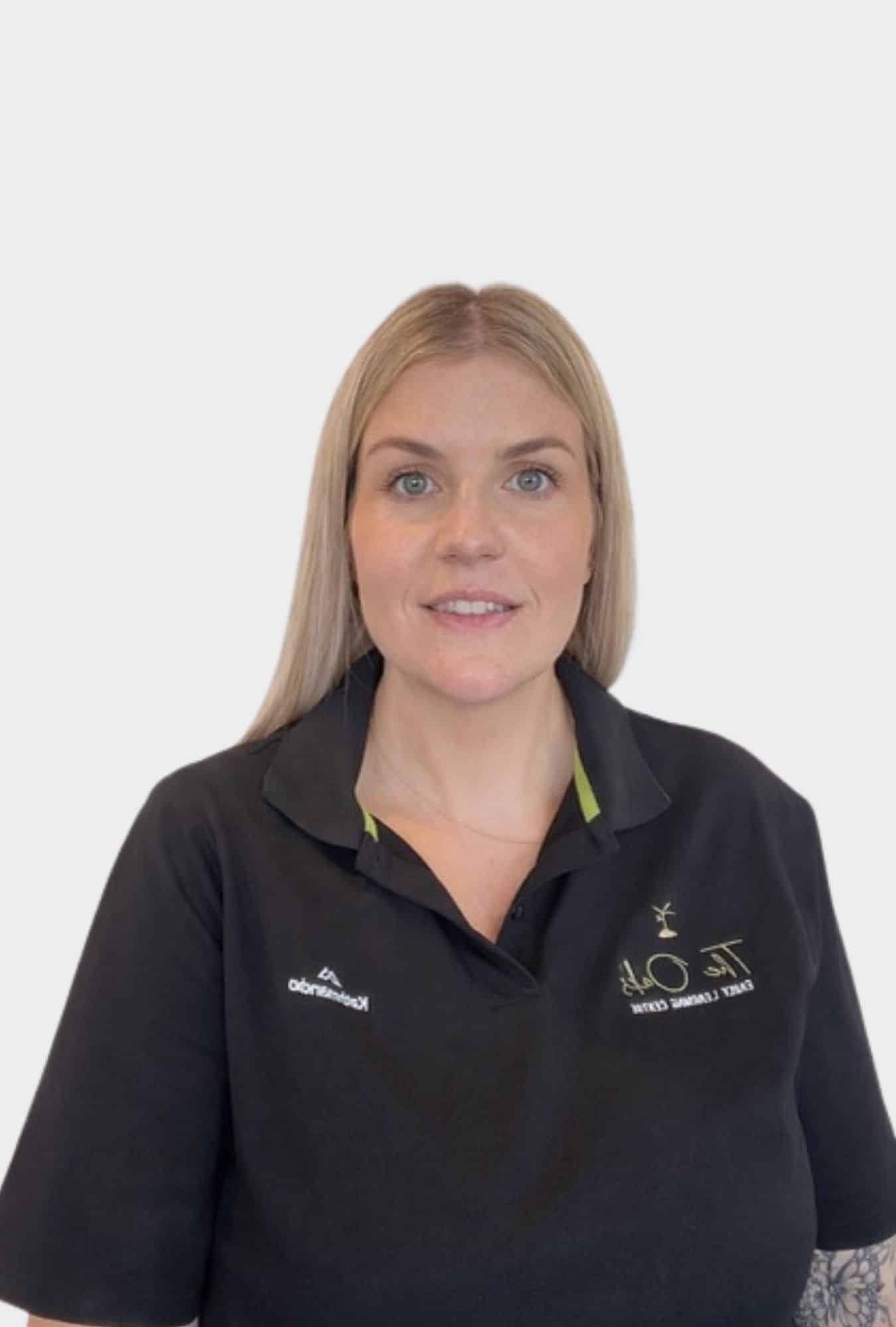 A woman with straight blonde hair is wearing a black collared shirt with embroidered logos, standing against a plain white background.