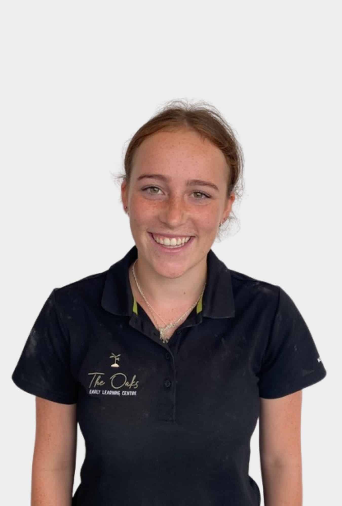 A young woman with brown hair smiles at the camera, wearing a navy blue polo shirt with "The Oaks Early Learning Centre" logo. She stands against a plain white background.