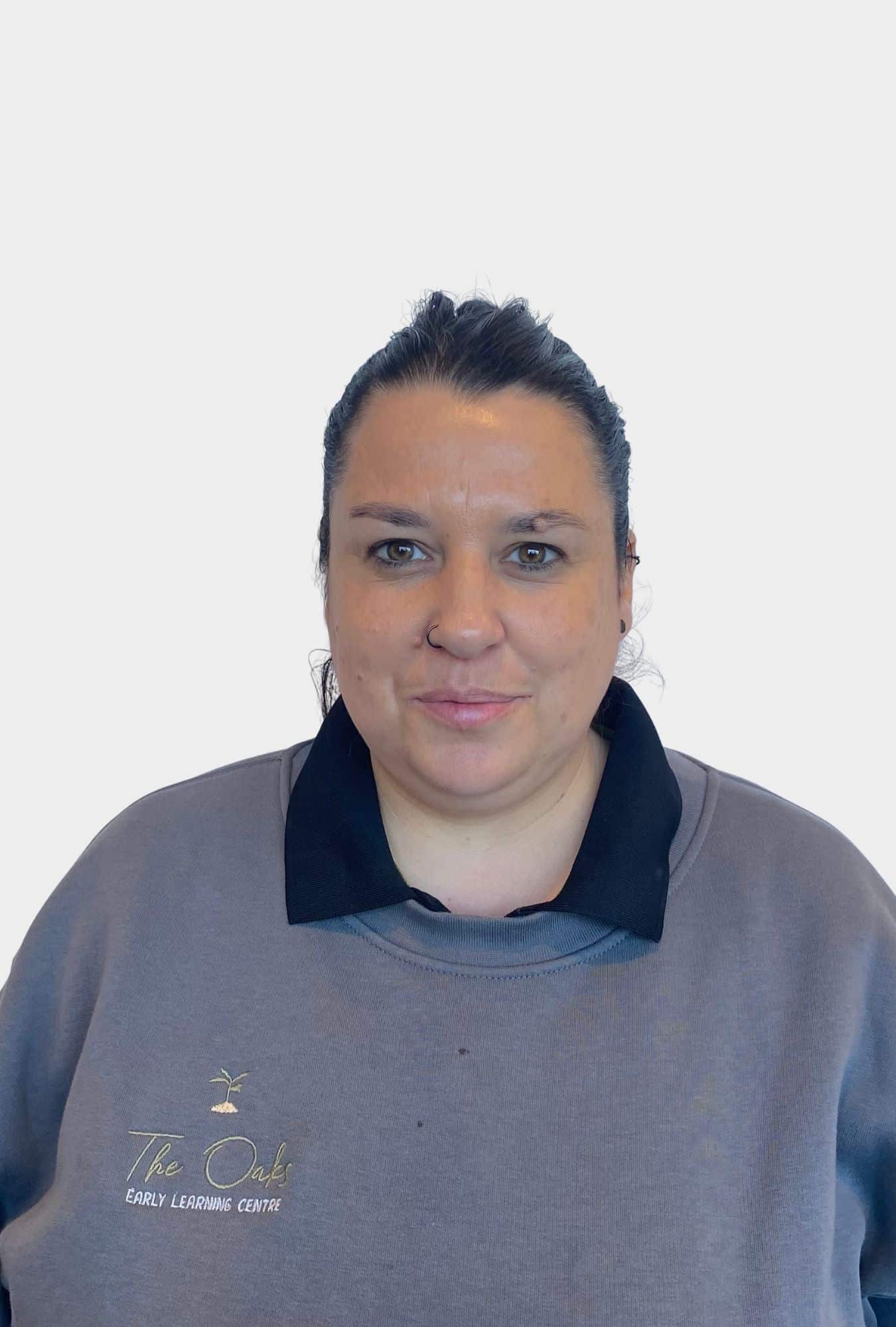 A woman with dark hair tied back, wearing a grey sweatshirt with "The Oaks Early Learning Centre" logo, stands against a plain white background.