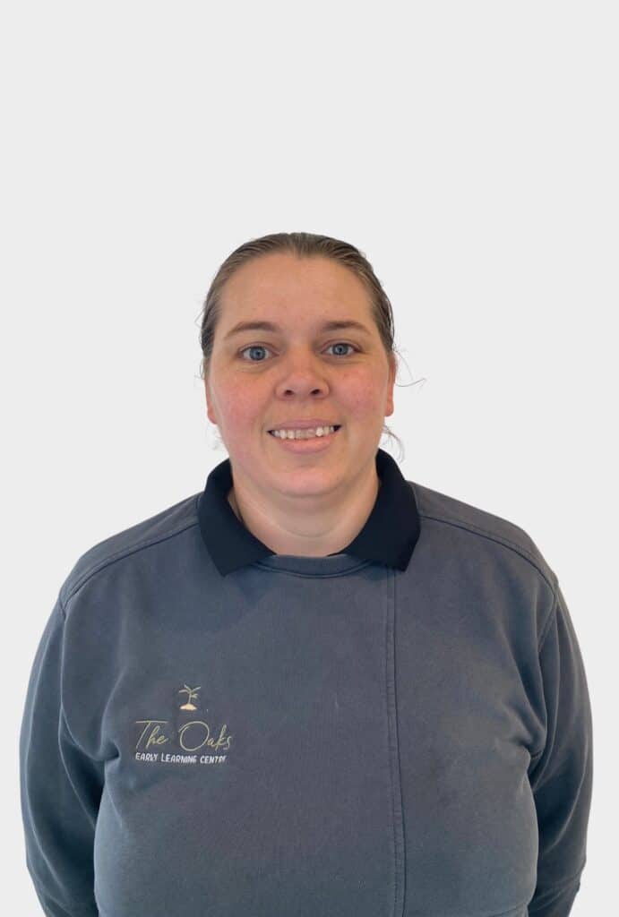 A person wearing a dark sweatshirt with "The Oaks Early Learning Centre" logo stands against a plain white background, smiling at the camera.