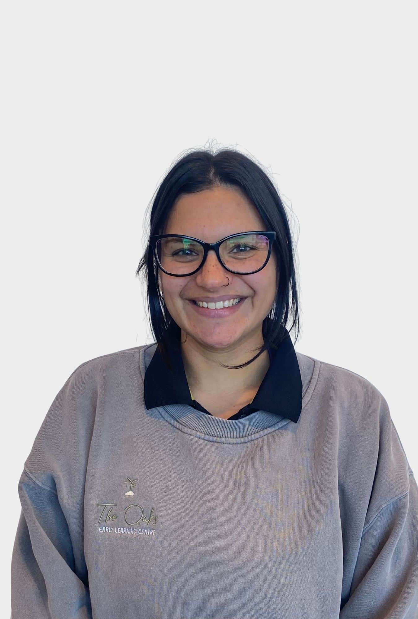A person with dark hair and glasses smiles, wearing a grey "The Oaks Early Learning Centre" sweatshirt over a black collared shirt, standing against a plain white background.