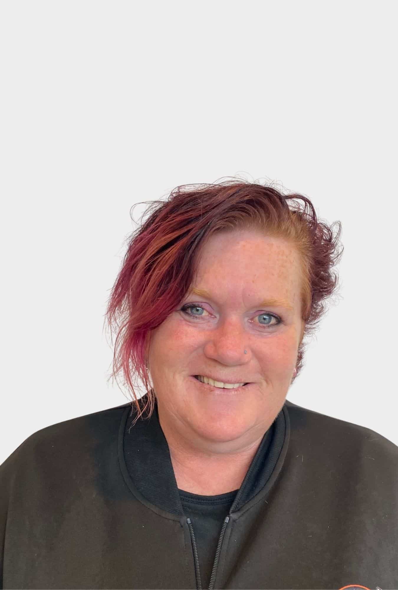 A woman with short, red hair and blue eyes is smiling at the camera, wearing a black jacket against a plain white background.